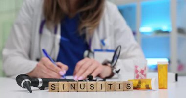 Tahta küplerde otoskop ve sarı hap şişesinin yanında Sinüzit yazıyor. Kadın doktor kronik sinüs ağrısı için reçeteli takip süreci yazıyor.