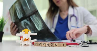 Kadın doktor, omurganın röntgen görüntüsünü dikkatlice inceler. Tahta küpler, hastanın teşhis konulmuş durumunu açıkça gösteren kelime kifozu oluşturur.