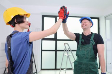 İki neşeli inşaat işçisi aynı beşliği paylaşıyor, gösterişli takım çalışması ve dostluk. Güvenlik kaskı ve eldiven takıyorlar, güzel ışıklandırılmış bir yerde anın tadını çıkarırken gülümsüyorlar..