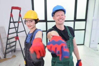 Renkli şapkalar ve eldivenler giyen iki hevesli inşaat işçisi, kapalı alanda iyi ışıklandırılmış bir alanda izleyiciye araçları gösterirken gülümsüyor.