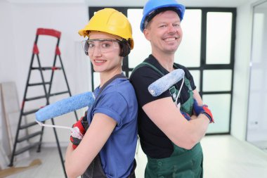 İki neşeli ressam sırt sırta dururlar, ellerinde boya rulolarıyla güvenle gülümserler. Parlak, ferah bir odada birlikte ev yenileme projesini yürütmeye hazırlar..