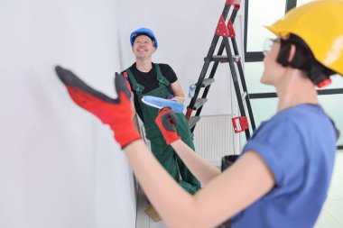 İki işçi ferah bir odada, biri merdivende, diğeri de yakınlarda boya aletleriyle meşguller. Duvarlar için yeni bir görünüm oluştururken gülümseme paylaşıyorlar..