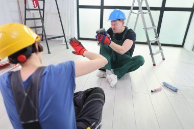 İki işçi aydınlık bir odada keyifli bir anı paylaşıyor. Biri bir matkabı neşeyle tutarken, diğeri yerde oturup el hareketleriyle işaret ediyor. Merdiven ve aletler etraflarını sardı..