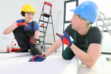Güneşli bir kapalı mekan ortamında, iki yetenekli müteahhit bir proje üzerinde çalışırken dostça bir değiş tokuş yapıyorlar. Şevklerini ve takım çalışmalarını gösteren sert şapkalar ve eldivenler giyiyorlar..