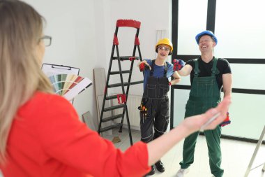 İki neşeli işçi, elinde renk örnekleri tutan bir kadınla, yenileme aletleri ve bir merdivenle meşgul oluyor. Oda doğal ışıkla parlıyor, canlı bir atmosfer yaratıyor..