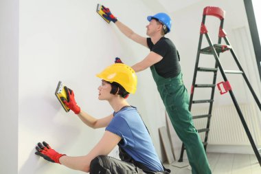 İyi ışıklandırılmış bir odada, iki işçi duvarlara yeni yapıştırılmış alçı yapıştırmaya odaklanıyor. Biri merdivenin üzerinde dururken diğeri çömelir, hem alet edevat hem de güvenlik teçhizatı ile donatılmıştır..