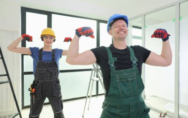 İki hevesli inşaat işçisi, yenilenmiş bir ofis alanında şakacı bir poz verirken kaslarını esnetiyorlar. Güneş ışığı geniş pencerelerden akar ve neşeli bir atmosfer yaratır..