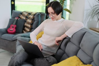 Doğal ışıkla dolu şık bir oturma odasında, dizüstü bilgisayarında yazan genç bir kadın, keyifli yastıklarla çevrili yumuşak gri bir kanepeye rahatça oturuyor ve arka planda bir gitar var..