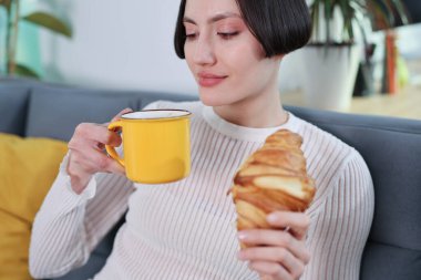 Sıcak bir fincan kahvenin ve gevrek bir kruvasanın tadını çıkaran bir kadın, parlak renkler ve bitkilerle çevrili bir kanepede rahatça oturur ve bir an için rahatlar..