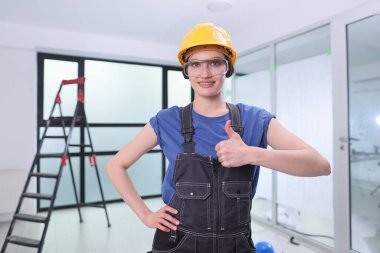 Sert şapkalı ve tulumlu kendine güvenen bir kadın, parlak bir inşaat alanında başparmağıyla poz veriyor. Ayarların bir merdiveni var ve potansiyel dolu, bu da ilerleme ve takım çalışması anlamına geliyor..