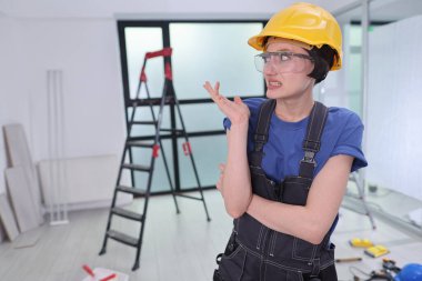 Sarı şapkalı ve gözlüklü bir inşaat işçisi alet ve merdiven dolu bir odada duruyor. Sinirli görünüyor, yenileme projesindeki bir sonraki hamlesini düşünüyor..