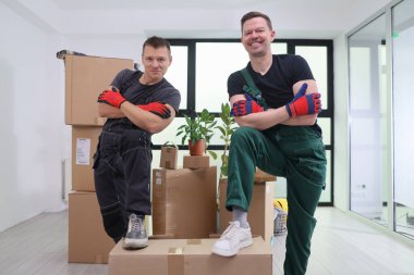 İki enerjik nakliyeci, çeşitli karton kutuların arasında kollarını kavuşturarak güvenle dururlar. Renkli eldivenler giyiyorlar ve gülümsüyorlar. Takım çalışmalarını güneşli ve modern bir mekanda sergiliyorlar..