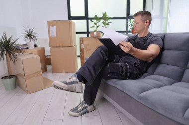 Günlük iş kıyafeti giymiş bir adam önemli belgeleri incelerken rahat bir koltukta dinleniyor. Yakınlarda karton kutular var. Yeni bir eve taşındıklarını ima ediyorlar..