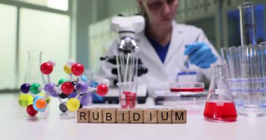 Tahta küpler parlak bilim laboratuarında rubidyum kelimesini heceler. Kadın bilim adamı, element davranışını açıklayan mikroskop ve renkli molekül modeli kullanır.