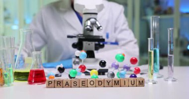 Tahta küpler Praseodiyum kelimesini temiz beyaz masa üzerinde heceler. Kadın doktor modern mikroskopla çalışan renkli moleküler modeller üzerinde çalışıyor.