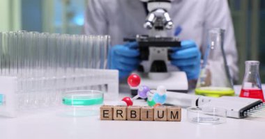 Tahta küpler, parlak klinik laboratuvarında Erbium kelimesini heceler. Kadın doktor mikroskop kullanarak test tüplerinin yanında detaylı moleküler deney hazırlıyor.