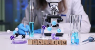 Tahta küpler Thallium kelimesini laboratuvar masasında cam eşyayla heceler. Kadın doktor mavi solüsyon ve mikroskop yakınında renkli moleküler modeller üzerinde çalışıyor.