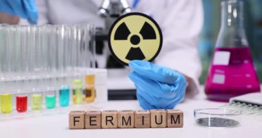 Tahta küpler, radyoaktif uyarı etiketinin yanındaki Fermium kelimesini heceler. Kadın doktor, araştırma laboratuarında test tüplerini düzenleyen renkli çözümlerle çalışıyor.