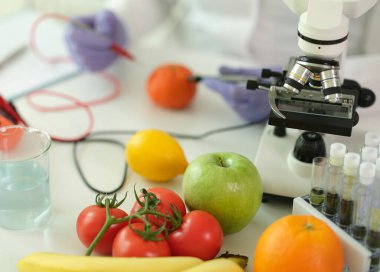 Parlak bir laboratuvarda, bir bilim adamı çeşitli meyveleri mikroskop altında inceler. Renkli domatesler, portakallar, limonlar ve yeşiller masayı dolduruyor, doğa ve bilimi harmanlıyor..