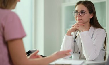 Modern bir klinik ortamında, beyaz önlüklü bir doktor, hastanın sağlık sorunlarını yakından dinlemektedir. Oda aydınlık ve sakin, güven verici bir atmosfer yaratıyor..