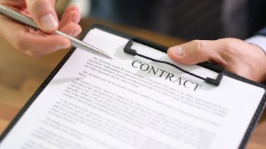 Bir iş adamı elinde bir kalem tutuyor, kağıt işleriyle çevrili bir panoda kontrat imzalamaya hazırlanıyor. Sıcak güneş ofis penceresinden parlıyor..