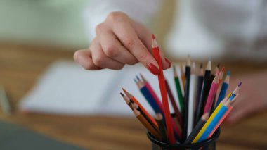 Parlak tırnak cilasıyla süslenmiş bir el çeşitli renklerle dolu bir bardaktan kırmızı renkli bir kalem seçer. Ortam rahat bir çalışma alanı, yaratıcılık yayıyor..