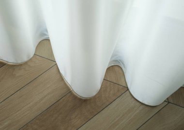 Narin beyaz perdeler taşar, yumuşak kıvrımları sıcak, parlak ahşap bir zemine düşer. Güneş ışığı bulutların arasından süzülür, sakin ve davetkar bir atmosfer yaratır..