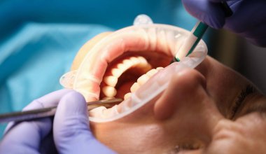 Diş hekimi özel aletler kullanarak dişlerini dikkatlice incelerken bir hasta dişçi koltuğuna yaslanır. Parlak klinik ortam, sakinlik duygusunu arttırır..