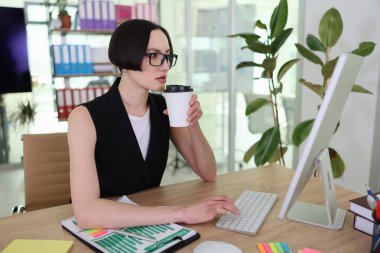 Kendini işine adamış bir birey, sıcak bir fincan kahve tutarken düzenli bir masada oturup klavyeye yazı yazar. Parlak ışık modern ofisi dolduruyor ve üretkenliğin canlı atmosferini arttırıyor..
