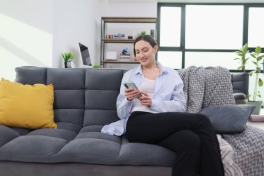 Rahat, genç bir kadın şık, gri bir koltukta, elinde telefonuyla rahatça oturur. Doğal ışık odayı dolduruyor, öğleden sonraki keyfi için sıcak ve davetkar bir atmosfer yaratıyor..