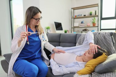 Beyaz önlüklü bir doktor koltukta dinlenen hamile bir kadınla nazikçe konuşur. Hasta endişeli görünüyor, alnını tutuyor. Doktor bu rahat evde sıcak bir yaklaşım sergiliyor..