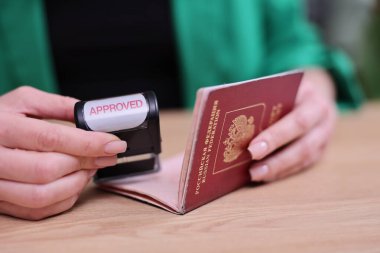 Elleri pasaport tutuyor, masanın üstünde duruyor. Onaylanmış bir pul bastırılır ve başlamak üzere olan bir yolculuğun habercisi olur. Bu an gelecek için umutla doludur..