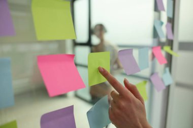 Bir el, cam bir duvardaki canlı pembe ve mor notalar arasında yeşil yapışkan bir nota işaret ediyor. Arka planda, bir insan düşünceli bir şekilde bakar, fikirler ve yaratıcılıkla çevrili..