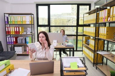 Hareketli bir ofis ortamında, bir kadın modern bir masada otururken belgelere göz atar, diğeri ise bilgisayara odaklanır. Parlak renkler ve doğal ışık odayı dolduruyor, verimliliği vurguluyor..