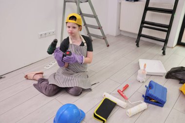Yerde oturan genç bir müteahhit, boya ruloları, bir merdiven ve çeşitli malzemelerle çevriliyken iki elektrikli aletle odaklanmış. Odanın yeni ve bitmemiş bir görünüşü var..