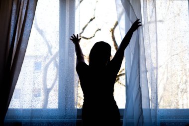 Bir kızın silueti bir pencerenin önünde sabah ışığıyla aydınlatılıyor.