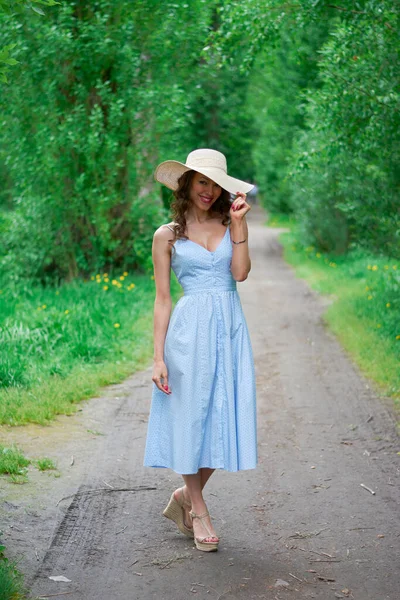 Όμορφο κορίτσι σε ένα ψάθινο καπέλο και μπλε φόρεμα στο δρόμο στο πάρκο — Φωτογραφία Αρχείου