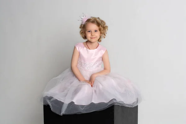 Una niña en un vestido rosa y corona se sienta en un cubo negro sobre un fondo blanco —  Fotos de Stock