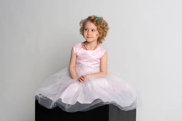 Una niña en un vestido rosa y corona se sienta en un cubo negro sobre un fondo blanco —  Fotos de Stock