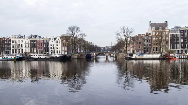 AMSTERDAM, PAÍSES BAJOS 31 DE MARZO DE 2016. Vista típica urbana en primavera. El río Amstel y los edificios de la construcción XVII-XVIII sobre terraplenes. Casas flotantes cerca de banco —  Fotos de Stock