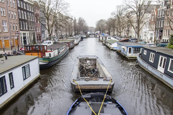 AMSTERDAM, NETHERLANDS on MARCH 31, 2016. Typical urban view. A row of traditional architecture on the canal embankment. The barge loaded with the sunk bicycles floats on the channel. Houseboats near the banks — Stock Photo, Image