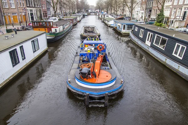 AMSTERDAM, NETHERLANDS on MARCH 31, 2016. Typical urban view. A row of traditional architecture on the canal embankment. The barge loaded with the sunk bicycles floats on the channel. Houseboats near the banks — Stock Photo, Image