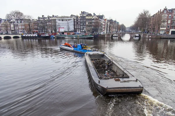 AMSTERDAM, NETHERLANDS on MARCH 31, 2016. Typical urban view. A row of traditional architecture on the canal embankment. The barge loaded with the sunk bicycles floats on the channel. Houseboats near the banks — Stock Photo, Image