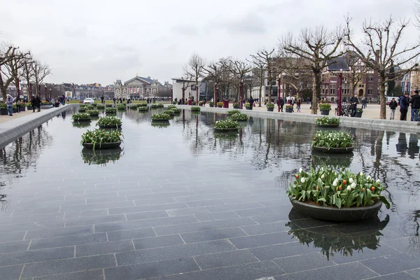 AMSTERDAM, PAÍSES BAJOS 31 DE MARZO DE 2016. Vista urbana. La fuente en la plaza del museo decorada con macetas con tulipanes. Los turistas se divierten . — Foto de Stock