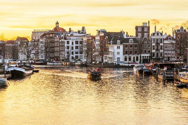 Amsterdam, Nederland op 31 maart 2016. Een typisch stedelijke uitzicht tijdens de zonsondergang. De Amstel rivier en de gebouwen van de bouw van de Xvii-Xviii op taluds. De wandelende schip drijft op de rivier — Stockfoto