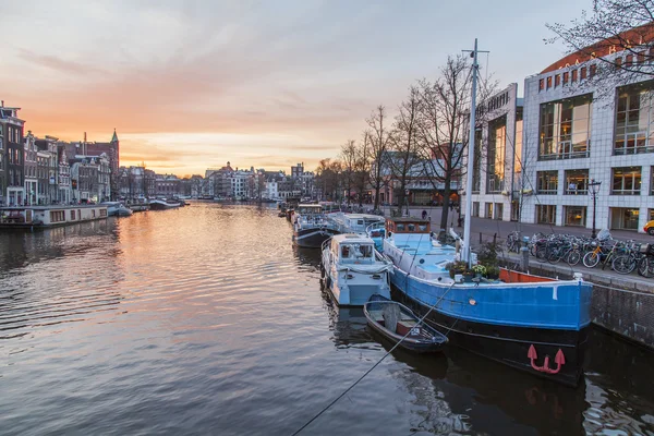 Amsterdam, Nederland op 31 maart 2016. Een typisch stedelijke uitzicht tijdens de zonsondergang. De Amstel rivier en de gebouwen van de bouw van de Xvii-Xviii op taluds. Vaartuigen die afgemeerd in de buurt van bank — Stockfoto