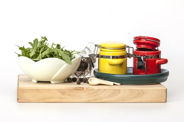 Zwarte peper in een transparante mortier met een stamper, bladeren van rucola en multi-gekleurde containers voor specerijen — Stockfoto