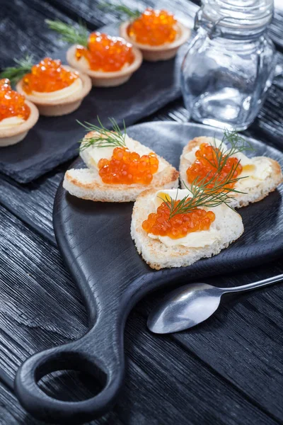Lanches em forma de coração com manteiga e caviar vermelho servido com endro em uma mesa de madeira preta — Fotografia de Stock