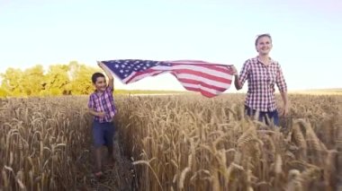 Little kid with mom holding an American flag on the wind in a field of wheat. Summer landscape against the blue sky.