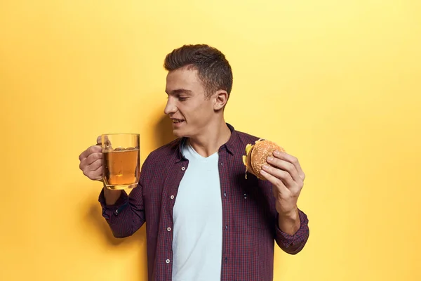 Homme joyeux avec une tasse de bière et un hamburger sur ses mains régime alimentaire style de vie jaune fond — Photo